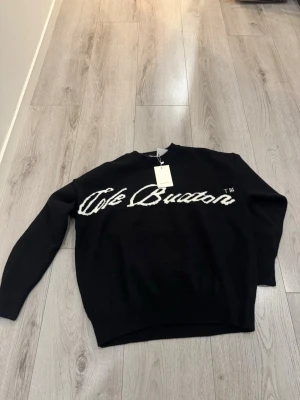 Svart stickad tröja Cole Buxton - Svart stickad tröja från Cole Buxton med vit broderad logga över bröstet. Tröjan har rund halsringning, långa ärmar och ribbade muddar vid ärmslut och nederkant. Perfekt för dig som gillar streetwear/archive och enkel stil.