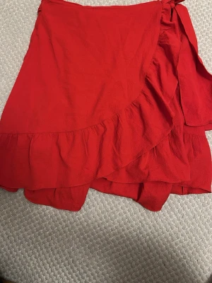 Röd omlottkjol med volang - Snygg röd kortkjol i omlottmodell med volangkant. Kjolen har knyt i sidan och är tillverkad i ett lätt, mjukt tyg som ger en skön känsla. Perfekt för dig som vill ha en färgstark och trendig look.