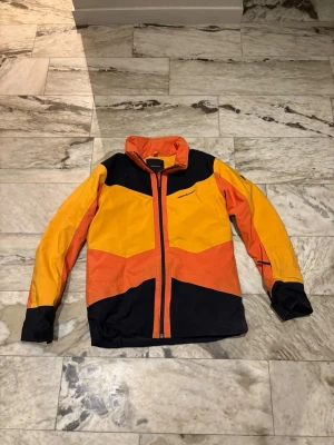 Färgstark skidjacka från peak performance - Säljer en riktigt snygg skidjacka från peak performance i orange, gul och svart. Jackan har hög krage, dragkedja framtill och coola färgblock. Tyvärr så saknar den huva. Passar perfekt på dig som är runt 170 till 175 cm lång.Tillverkad i slitstark polyester och perfekt för dig som vill sticka ut. Passar till både stad och äventyr! 