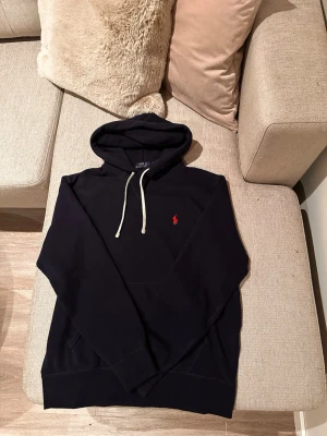 Mörkblå hoodie från Polo Ralph Lauren - Snygg mörkblå hoodie från Polo Ralph Lauren med klassisk röd logga broderad på bröstet. Hoodien har vit dragsko i huvan och en stor magficka framtill. Perfekt för en avslappnad och stilren look. Helt oanvänd!