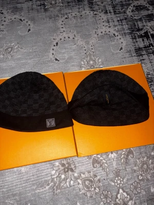 Svart Louis Vuitton mössa - Snygg svart mössa från Louis Vuitton med diskret rutmönster och LV-logga framtill. Tillverkad i mjukt material som känns lyxigt och bekvämt. Perfekt accessoar för dig som vill ha en exklusiv touch på din outfit.