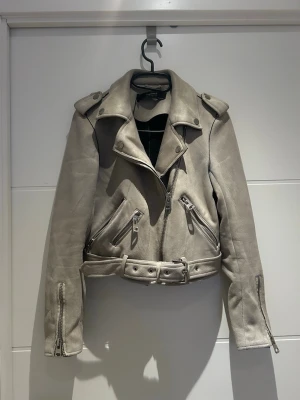 Beige mockajacka med bälte från Zara - Snygg beige skinnjacka från Zara med coola detaljer som bälte i midjan, dragkedjor och axelklaffar. Jackan har en croppad passform och flera fickor med dragkedja. Perfekt för dig som vill ha en edgy stil.