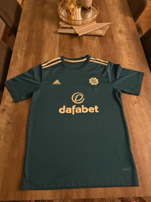 Grön Adidas Celtic matchtröja - Snygg grön matchtröja från Adidas med Celtic FC-logga och guldfärgade detaljer. Tröjan har korta ärmar, rund hals och tre guldiga ränder på axlarna. Tillverkad i lätt och ventilerande polyester, perfekt för fotboll eller träning. Lite sönder men de syns knappt 