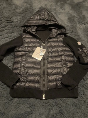 Svart dunjacka med huva från Moncler - Svart dunjacka från Moncler med quiltad kropp, stickade ärmar och huva. Jackan har dragkedja framtill, ribbade muddar och en ficka med dragkedja på ena ärmen. Perfekt för kalla dagar och riktigt snygg streetstil.