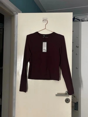 Mörkröd långärmad topp från Pull&Bear - En stilren mörkröd långärmad topp från Pull&Bear. Modellen är croppad med rund halsringning och har en enkel, clean look utan mönster eller detaljer. Perfekt att matcha med höga jeans eller kjol. Materialet är mjukt och stretchigt, tillverkat i bomull och elastan. 