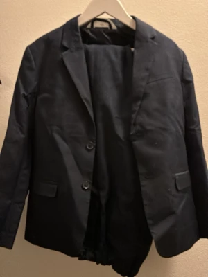 Mörkblå kostym, kavaj med byxor barn storlek 140 - Säljer en klassisk mörkblå kostym med kavaj och matchande byxor. Kavajen har två knappar framtill, smala slag och fickor med lock. 