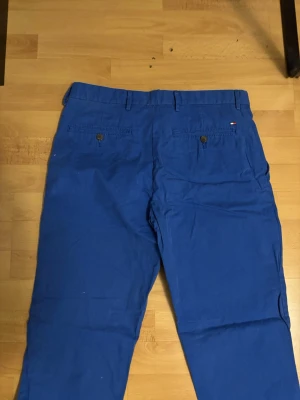 Blå chinos från Tommy Hilfiger - Snygga blå chinos från Tommy Hilfiger med raka ben och klassisk passform. Byxorna har bälteshällor, två fickor fram och två bakfickor med knapp. Perfekta för en stilren och avslappnad look.