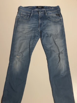Replay Hyperflex Anbass ljusblå jeans - Säljer ett par Replay Hyperflex Anbass jeans i ljusblå tvätt med slitningar och trasiga detaljer på benen. Klassisk femficksmodell med dragkedja och smal passform. Superstretchigt material som är riktigt skönt att bära.