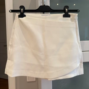 Vita skorts med omlottdetalj, Stradivarius  - Stilrena vita skorts med omlottdetalj framtill och diskret dragkedja i sidan. Klassisk och enkel design som passar perfekt till sommaren. Tillverkade i ett mjukt tyg som ger en bekväm känsla och snygg passform.