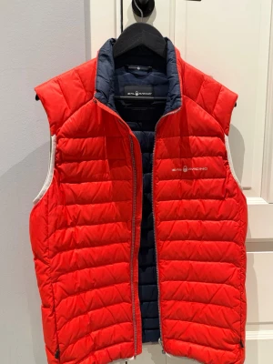 Röd dunväst Sail Racing - Snygg röd dunväst från Sail Racing med marinblått innerfoder och diskret logga på bröstet. Västen är herr, i storlek L. Köpt på Sail racing butik inne i Göteborg. 