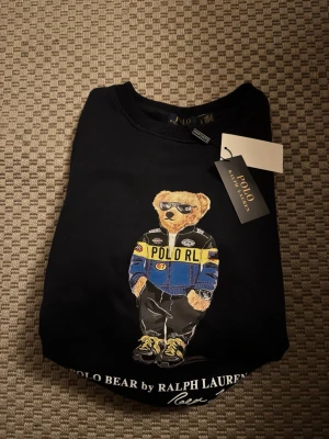 Svart Polo Bear sweatshirt Ralph Lauren storlek s - Svart sweatshirt från Ralph Lauren med ikoniska Polo Bear-trycket framtill. Björnen har på sig en blå och gul jacka med solglasögon. Tröjan har rund hals, ribbade muddar och är i mjukt material. Perfekt för en avslappnad och trendig look.