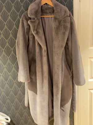 Grå lång pälsjacka med krage - Snygg och fluffig grå pälsjacka med bred krage och knappar framtill. Jackan är lång och har en oversized passform som ger en cool och trendig look. Perfekt för kalla dagar när du vill vara både varm och stilren.