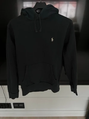 Svart hoodie från Polo Ralph Lauren - Klassisk svart hoodie från Polo Ralph Lauren med broderad logga på bröstet. Hoodien har huva med dragsko och en stor magficka framtill. Tillverkad i mjukt material som känns skönt mot huden. Perfekt för en avslappnad och stilren look.