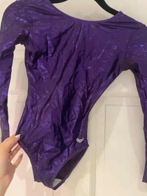 Lila glittrig body från Agiva - Säljer min gamla träningsdräkt från Agiva anpassad för AG eller truppgymnastik. Tillverkad i stretchigt tyg. Nypris: 699 kr. Storlek 12