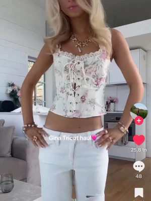 Blommig spetsbody från Gina Tricot - Supersöt body från Gina Tricot i vitt med blommigt mönster i rosa och grönt. Modellen har snörning framtill och spetskant vid urringningen. Perfekt för dig som gillar romantisk och trendig stil.