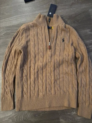 Beige kabelstickad tröja Polo Ralph Lauren - Kabelstickad beige tröja från Polo Ralph Lauren med halv dragkedja och hög krage. Tröjan har ribbade muddar och nederkant samt den klassiska mörkblå logotypen broderad på bröstet. Perfekt för lager på lager och snygg till jeans.