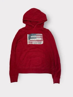 Röd hoodie från Ralph Lauren - Röd hoodie från Ralph Lauren med stor amerikansk flagga och texten 'Denim & Supply' på bröstet. Klassisk känguruficka framtill och huva. Tillverkad i mjuk bomull, perfekt för en avslappnad streetwear-look.