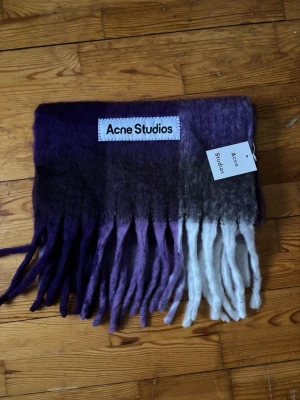 Lila-grå Acne Studios halsduk - Säljer en fluffig halsduk från Acne Studios i lila, grå och vit färgskala. Halsduken har långa fransar och stor Acne Studios-logga. Materialet är mjukt och borstat, perfekt för att ge din outfit en cool touch.