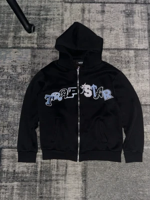 Svart Trapstar hoodie med dragkedja - Säljer en svart hoodie från Trapstar med stor huva och dragkedja framtill. Framsidan har ett stort Trapstar-tryck i blått, vitt och grått. Hoodien har två fickor och ribbade muddar vid ärmar och nederkant. Perfekt för en streetwear-look.
