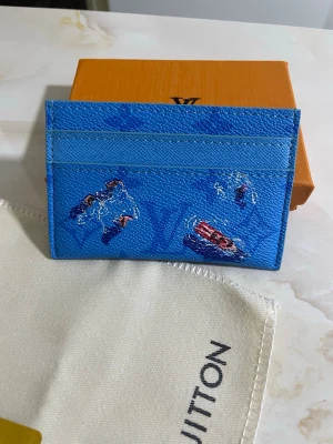 Blå korthållare - Säljer en blå korthållare med ett unikt tryck av badande personer och en röd båt. Plånboken har flera kortfack och är tillverkad i tåligt skinn med en lyxig känsla. Perfekt för dig som vill ha något färgglatt och exklusivt i fickan.