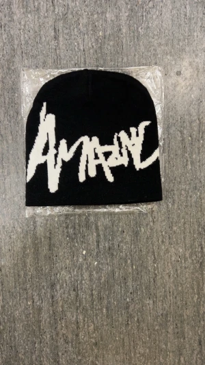 Svart beanie med vit Amaze-logga - Svart beanie med stor vit Amaze-logga i graffiti-stil framtill. Mössan är slätstickad och har en klassisk, rundad form. Perfekt för dig som gillar streetwear och vill sticka ut med en cool accessoar.