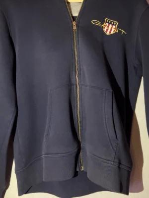 Mörkblå zip-hoodie från GANT - Mörkblå hoodie från GANT med dragkedja framtill och två stora fickor. Bröstet pryds av ett broderat GANT-emblem med texten 1949 i gult och rött. Klassisk college-look och ribbade muddar vid ärmslut och nederkant. Perfekt för en avslappnad stil.