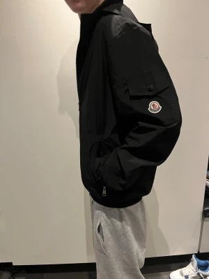 Svart Moncler vindjacka med fickor - Snygg svart vindjacka från Moncler med dragkedja framtill, hög krage och ficka med Moncler-logga på ärmen. Jackan har en clean look, perfekt för vår och höst. Passformen är normal och jackan har långa ärmar. Det är en 🐄Pia och den går att scanna den passar ungefär 170cm och neråt 