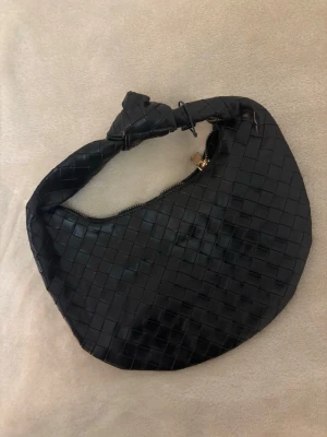 Svart flätad handväska med knut - Trendig svart handväska i flätad design med rundad form och en snygg knut på handtaget. Väskan har dragkedja i guld och är tillverkad i skinnimitation. Perfekt accessoar för att lyfta din outfit.