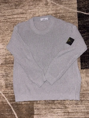 Grå stickad tröja från Stone Island - Säljer en grå stickad tröja från Stone Island med klassisk patch på ärmen. Tröjan har rund halsringning, ribbade muddar och en clean, oversized passform. Perfekt för dig som gillar streetwear och vill ha något stilrent men ändå fett.
