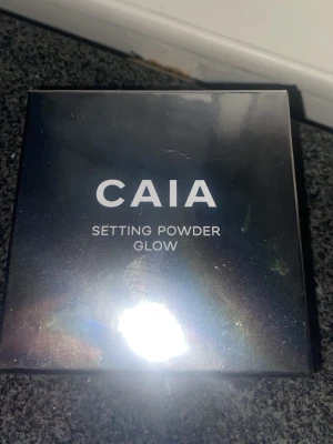 CAIA Setting Powder Glow - Säljer ett CAIA Setting Powder Glow som ger en snygg glow-effekt till din makeup. Kommer i en stilren, svart kompakt förpackning med spegeleffekt. Perfekt för att fixera sminket och få en fräsch finish.