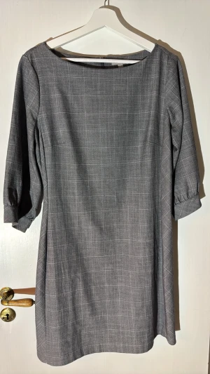 Grå rutig klänning från H&M - Stilren grå klänning från H&M med rutigt mönster och trekvartsärm. Klänningen har en rak passform och dragkedja i ryggen. Perfekt för dig som gillar enkel men snygg design.
