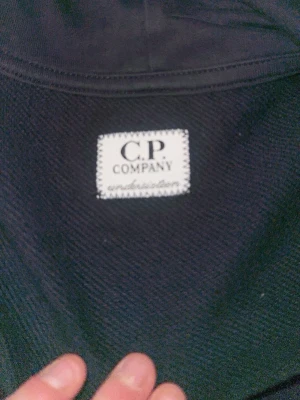 Svart hoodie från C.P. Company - Snygg svart hoodie från C.P. Company med dragkedja framtill och klassisk logga på bröstet. Hoodien har känguruficka och justerbar huva. Perfekt för en avslappnad streetwear-look.