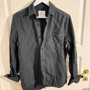 Mörkgrå manchesterskjorta A Day's March - Säljer en snygg mörkgrå manchesterskjorta från A Day's March. Skjortan har klassisk krage, bröstficka och knappar framtill. Perfekt för dig som gillar en stilren och avslappnad look. Passar till det mesta och är riktigt skön att bära.