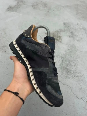Valentino Rockrunner sneakers svart - Säljer ett par svarta Valentino Rockrunner sneakers med coola nitar runt sulan och detaljer i mocka och skinn. Skorna har kamouflagemönster, snörning och en chunky sula som ger en edgy vibe. Perfekta för dig som vill sticka ut med stil. En nit saknas på baksidan