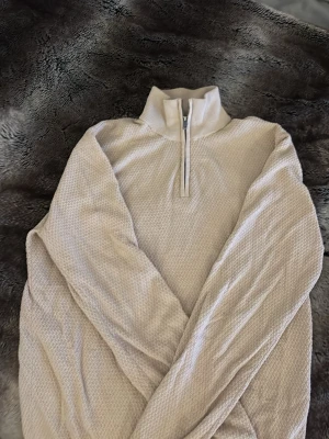 Beige stickad tröja med halv dragkedja - Säljer en beige stickad tröja från Dressmann med halv dragkedja och hög krage. Tröjan har ett diskret strukturmönster och ribbstickad krage. Perfekt för lager på lager och en clean look.
