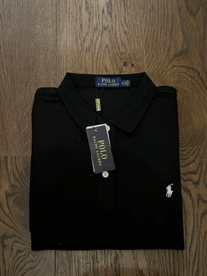 Svart pikétröja från Polo Ralph Lauren - Klassisk svart pikétröja från Polo Ralph Lauren med vit broderad logga på bröstet. Tröjan har krage, knappar framtill och är tillverkad i mjuk bomull som känns skön mot huden. Perfekt för en clean och stilren look.