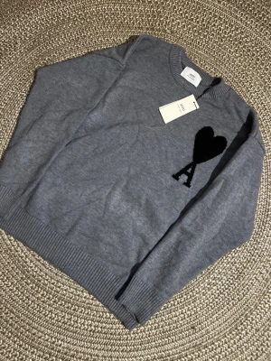 Grå stickad tröja från Ami - Snygg grå stickad tröja från Ami med svart hjärta och bokstaven A broderat på bröstet. Tröjan har rund halsringning, ribbade muddar och är tillverkad i mjukt material som känns lyxigt. Perfekt för dig som gillar stilrena detaljer.