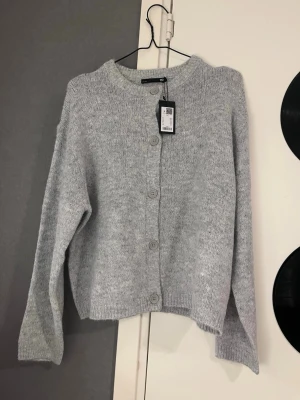 Ljusgrå kofta med knappar - Mysig ljusgrå kofta från lager157 med rund halsringning och stora matchande knappar framtill. Koftan är stickad i mjukt material och har långa ärmar samt ribbade muddar vid ärmslut och nederkant. Perfekt för lager på lager och enkel att styla.