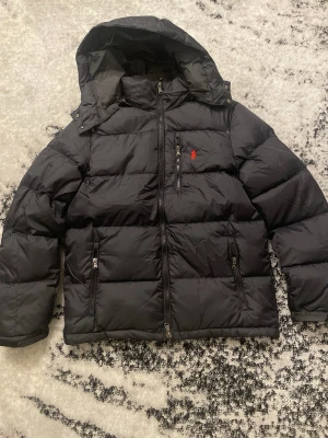 Svart pufferjacka från Polo Ralph Lauren - Säljer en svart pufferjacka från Polo Ralph Lauren med huva och dragkedjor. Jackan har det klassiska röda Polo-logot på bröstet och flera praktiska fickor med dragkedja. Perfekt för kalla dagar och riktigt snygg till streetwear-stilen.