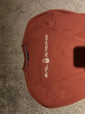 Rostbrun sweatshirt från Sail Racing - Säljer en snygg rostbrun sweatshirt från Sail Racing med vit logga tryckt på bröstet. Tröjan har rund halsringning och är tillverkad i mjukt bomullsmaterial. Perfekt för en avslappnad och sportig stil.