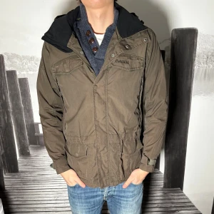 Brun parkasjacka från Berghaus - Snygg brun parkasjacka från Berghaus med huva och flera praktiska fickor framtill. Jackan har dragkedja och knappar, samt justerbar midja för bättre passform. Perfekt för kyligare dagar och har en klassisk outdoor-look.