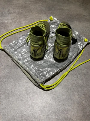 Nike fotbollsskor i neongult - Säljer ett par grymma Nike fotbollsskor i neongult med svart swoosh. Skorna har hög strumpliknande krage, snörning och är tillverkade i syntetmaterial med stickad känsla. Perfekt för dig som vill sticka ut på planen! Levereras med matchande Nike-påse.
