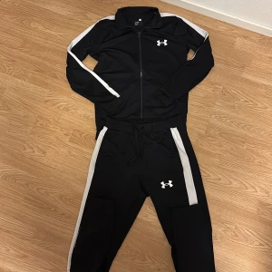 Under Armour tracksuit - Svart tracksuit från Under Armour med vita sidopaneler och logga på benet. Byxorna har snörning i midjan och är gjorda i ett mjukt syntetmaterial som passar perfekt för träning. Snygg sportig look med tapered passform.