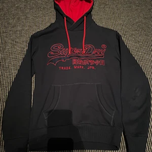 Blå Superdry Hoodie - En navy blue Superdry hoodie i väldigt bra skick och är enbart använd enstaka gånger. Är blå med röda detaljer med skönt och välgjort material med en cool och slick design.