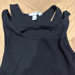 Svart ribbad linnetopp från H&M - Säljer en svart ribbad linnetopp från H&M i storlek S. Toppen har en cool cut-out detalj vid halsen och är ärmlös med rundad halsringning. Materialet är mjukt och stretchigt, perfekt för att skapa en trendig look. Passar dig som gillar minimalistisk stil.