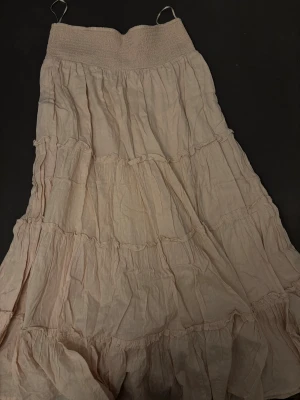 Beige volangkjol i bomull - Lång, ljusbeige maxikjol med flera volanger och smock i midjan. Kjolen är tillverkad i tunn bomull med diskret strukturmönster och har en luftig, flowy känsla. Perfekt för dig som gillar en bohemisk och avslappnad stil.