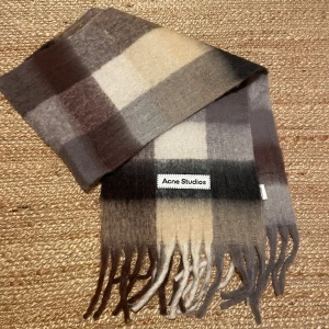 Rutig halsduk Acne Studios  - Snygg rutig halsduk från Acne Studios med stora blockrutor i beige, grått, brunt och svart. Mjuk och fluffig känsla i ullblandning, med långa fransar i ändarna och tydlig logotyp framtill. Perfekt nu till vintern för att ge din outfit en cool touch. Använd förra året men i mycket bra skick. Priset går att diskuteras. Nypris:3100