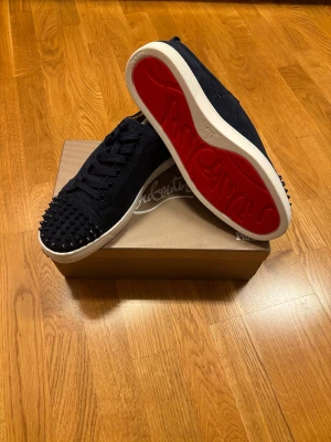 Louboutin sneakers mörkblå med spikar - Säljer ett par mörkblåa sneakers från Christian Louboutin med mocka-yttermaterial och coola mörkblåa nitar på tån. Skorna har snörning, vit sula och den ikoniska röda undersidan. Perfekt för dig som vill sticka ut med en edgy och lyxig stil.