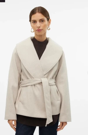 Beige omlott kappa med bälte - Säljer en beige kappa med bred krage och omlott-design. Jackan har ett matchande bälte i midjan och två sidofickor. Materialet känns mjukt och har en ull-liknande look. Perfekt för dig som gillar stilrena och chill plagg.