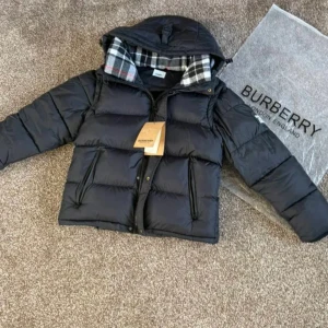 Svart pufferjacka från Burberry - Säljer en svart pufferjacka från Burberry med huva och dragkedja. Jackan har ett stort Burberry London England-märke på ärmen och klassiskt rutigt foder i huvan. Perfekt för kalla dagar och har en snygg, modern look.